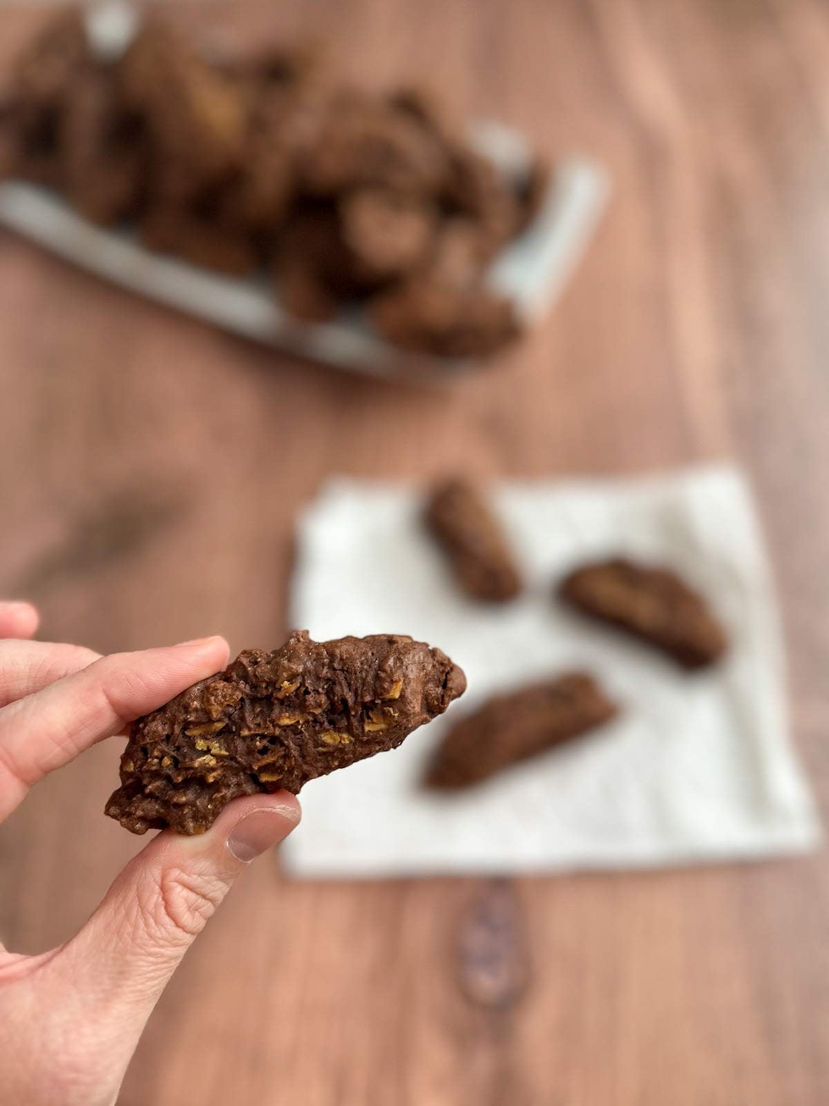 A hand holds a rectangular, crunchy, medium brown chocolate cookie with toasted almonds above a large plate FULL of other chocolate, biscotti-shaped chocolate pizzettes in this Italian Chocolate cookies recipe.