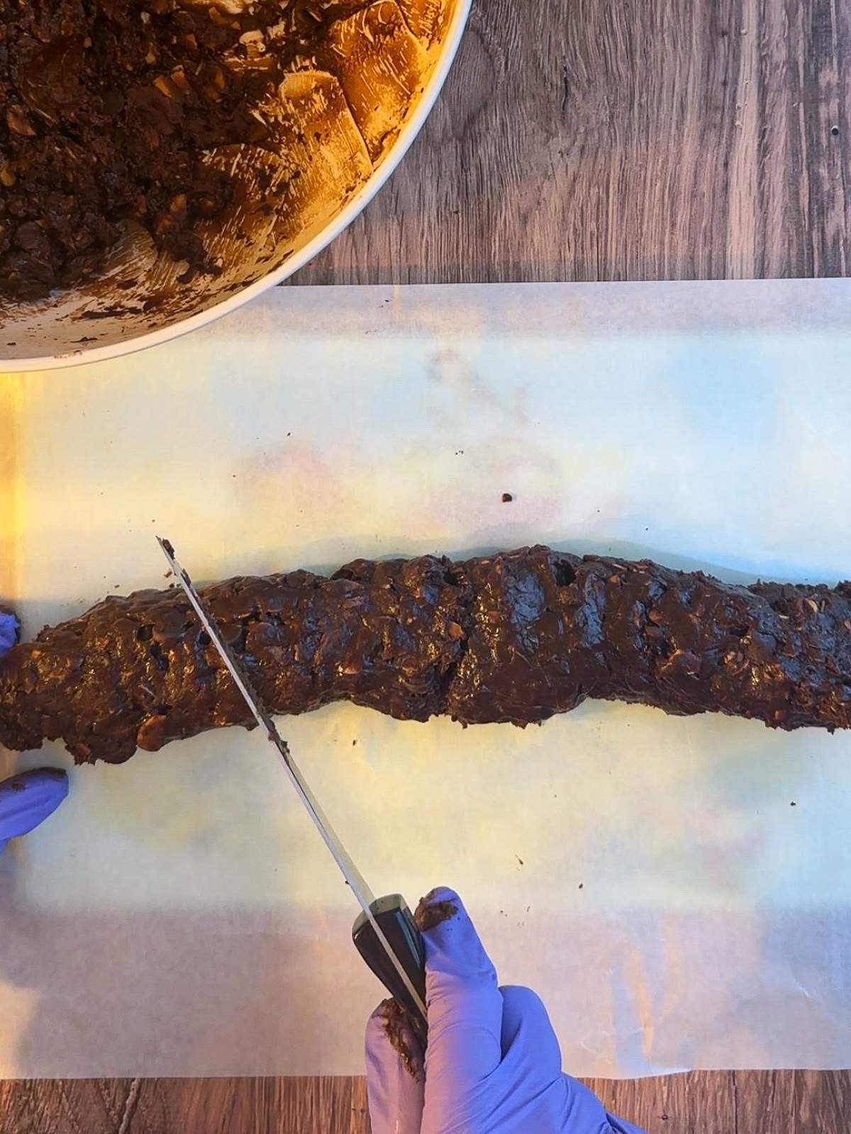 A gloved hand holds a knife diagonally to a log of chocolate dough on a piece of white parchment paper, on a cutting board, with a big bowl of more chocolate cookie dough with toasted almond pieces.