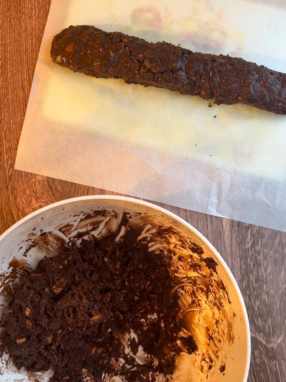 One log of dark brown chocolate cookie dough sits on a sheet of parchment paper on a cutting board, next to a large bowl of the remaining dark brown, chocolate cookie dough with toasted almond slices that hasn't been formed into cookie dough logs yet.