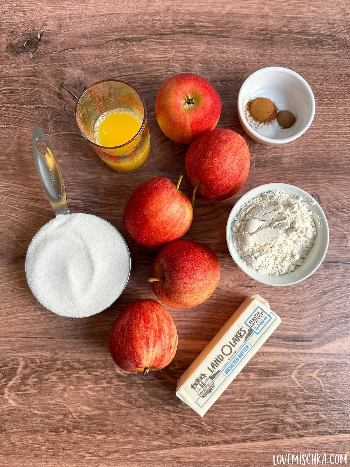 The simple ingredients of this classic Apple Betty Pie recipe - 5 bright red apples, a measuring cup of white granulated sugar, a small glass of orange juice, a small white bowl of two brown spices - cinnamon and nutmeg - and white salt, a cup of white flour, and a stick of cold unsalted butter.