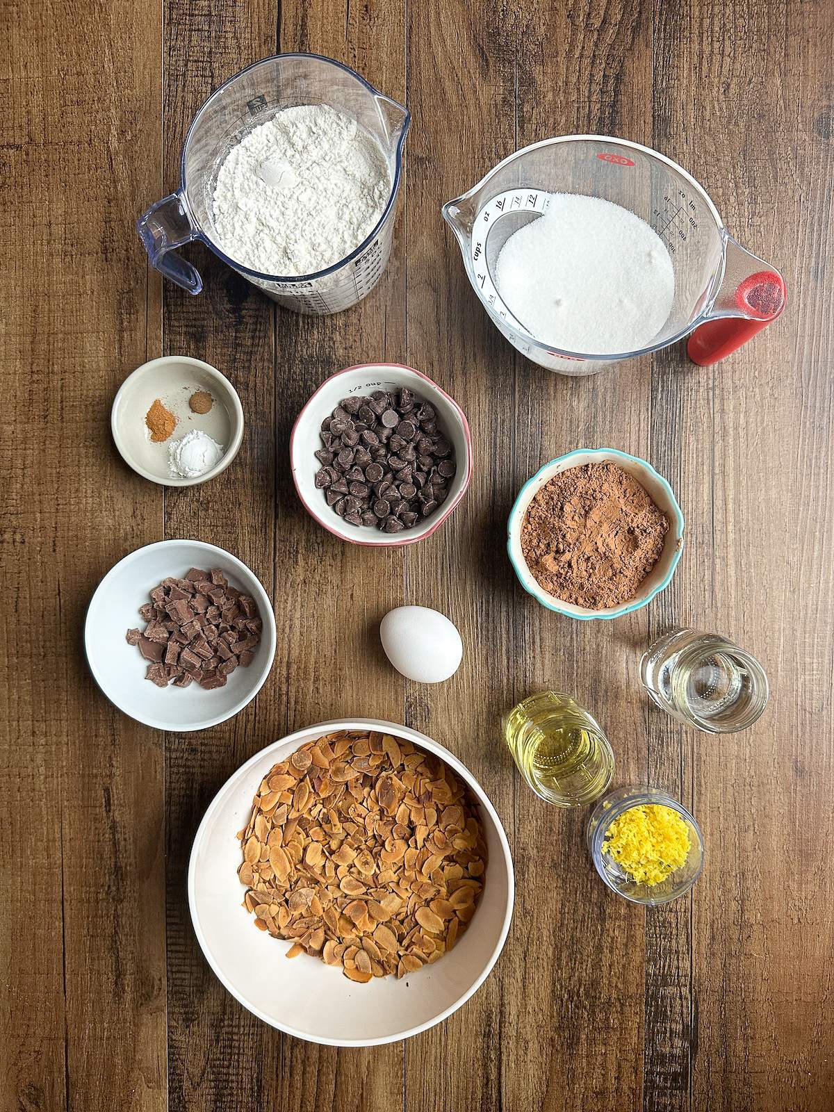 The ingredients in this Chocolate Italian Cookie Recipe, including semi-sweet chocolate chips, pieces of a Hershey's chocolate bar, Hershey's cocoa powder, lemon zest, an egg, water, olive oil, flour, sugar, baking powder, cinnamon, nut meg, and a large bowl of toasted almond slices. 