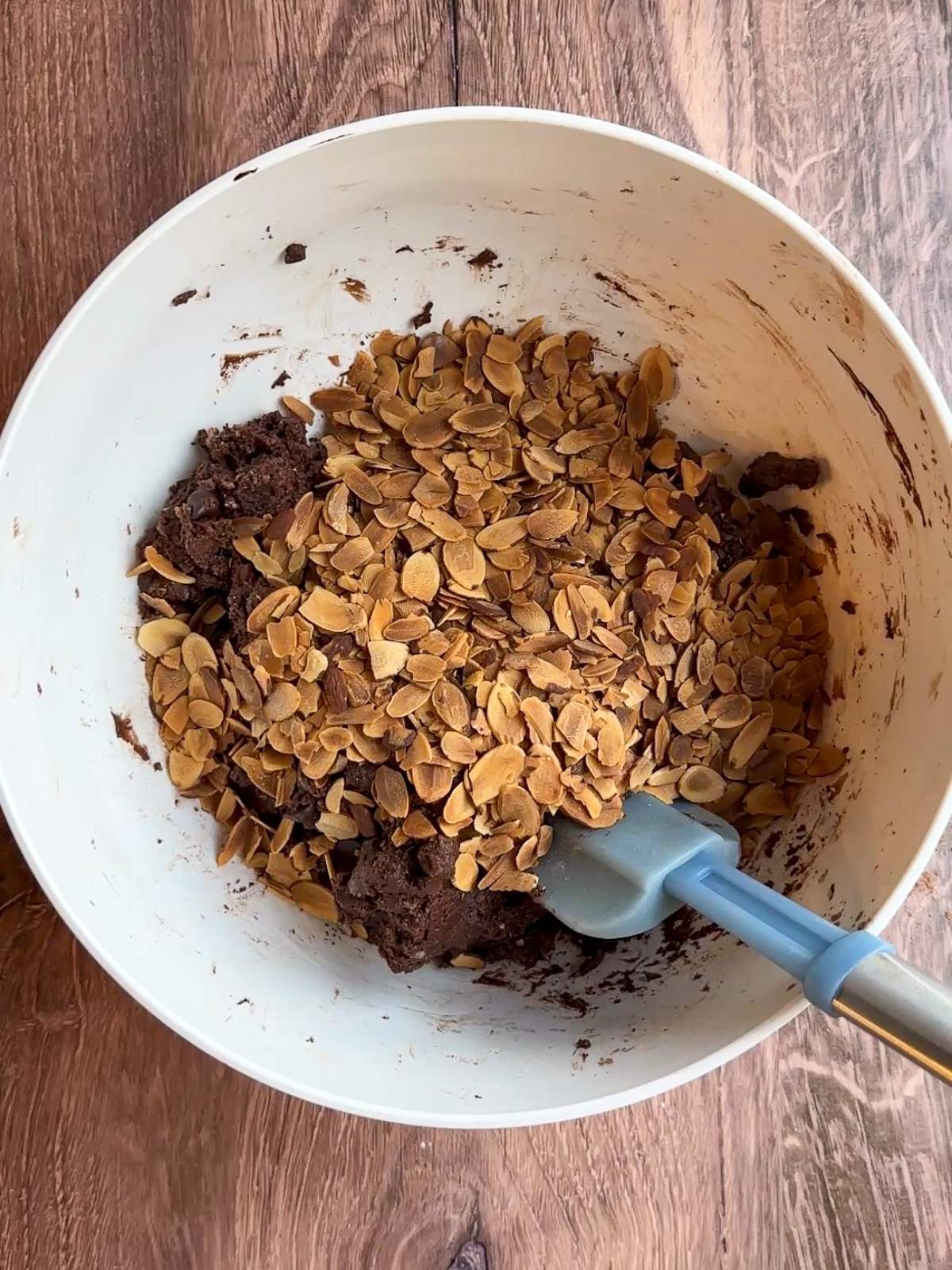 Golden brown toasted almond slices on top of recently mixed chocolate dough in a big, white bowl with a blue spatula in this Italian Chocolate Cookie Recipe. 