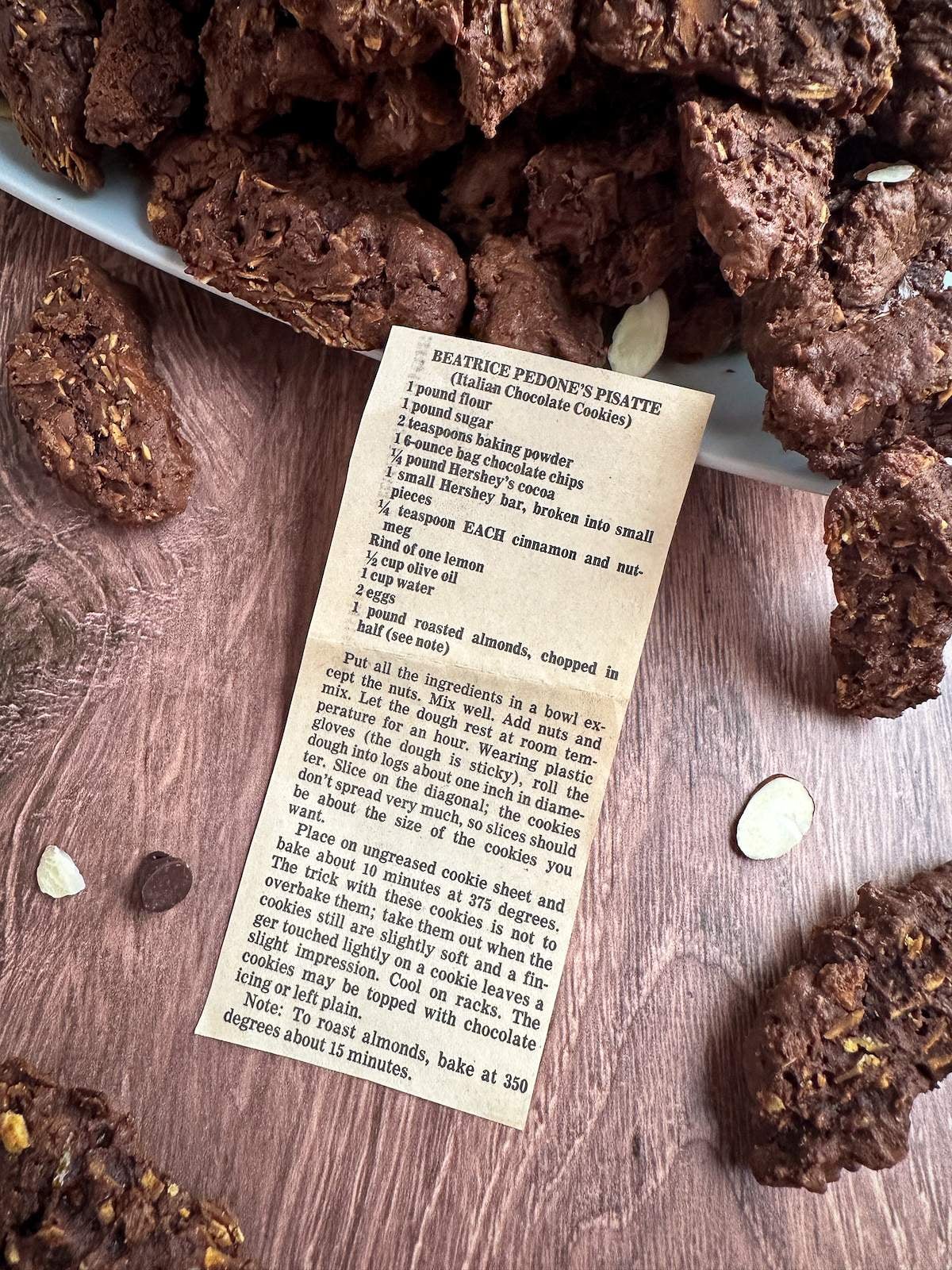 An old newspaper clipping of this chocolate Italian cookies recipe that says, "Beatrice Pedone's Pisatte (Italian Chocolate Cookies)" at the top. The newspaper clipping is surrounded by several, rectangular-shaped, chocolate cookies with crunchy toasted almonds inside, loose chocolate chips, and slices of raw almonds. 
