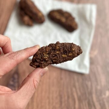 A hand holds an oval shaped, brown Italian chocolate cookie with toasted, tan almond slices above other biscotti-shaped chocolate Italian cookies on a folded sheet of white parchment paper.
