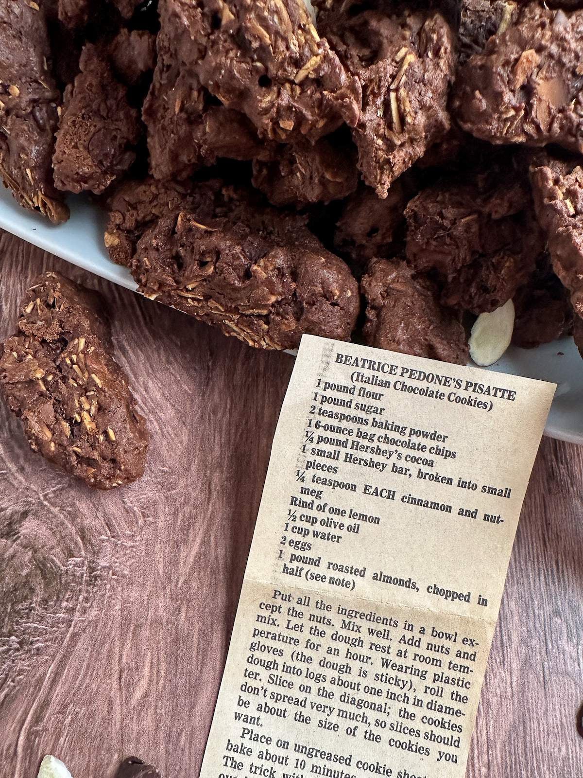 An old newspaper clipping for "Beatrice Pedone's Pisatte (Italian Chocolate Cookies)" recipe sits among brown, crunchy yet soft, chocolate cookies with almond pieces. Semi-sweet chocolate chips and raw almond slices are scattered amongst the cookies in this Italian cookie recipe.