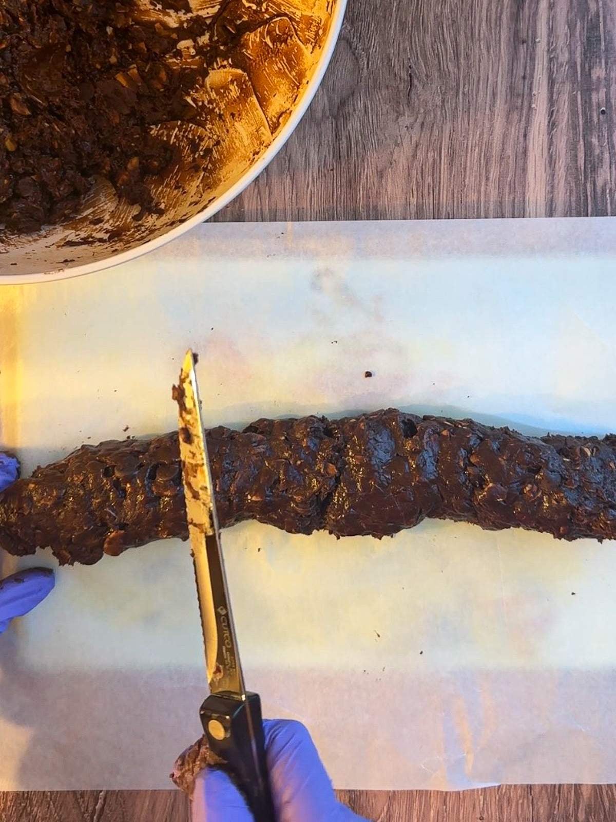 A gloved hand holds a knife at 45 degrees over a log of chocolate cookie dough in this chocolate Italian cookies recipe.